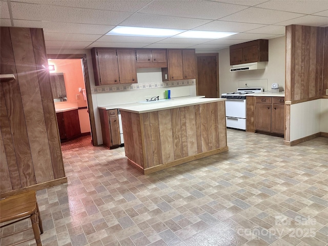 kitchen with white gas stove, a drop ceiling, light countertops, baseboards, and under cabinet range hood