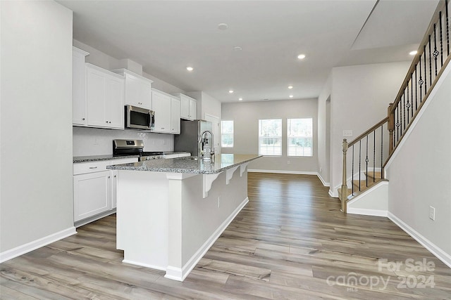 kitchen with dark stone countertops, an island with sink, light hardwood / wood-style floors, white cabinets, and appliances with stainless steel finishes