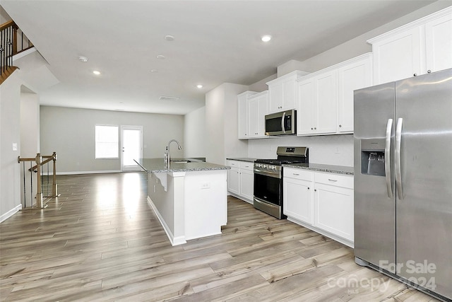 kitchen with appliances with stainless steel finishes, a kitchen island with sink, dark stone countertops, white cabinets, and light hardwood / wood-style floors