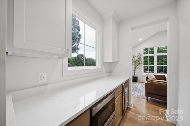 bar with stainless steel microwave, light stone counters, white cabinets, vaulted ceiling, and light wood-type flooring