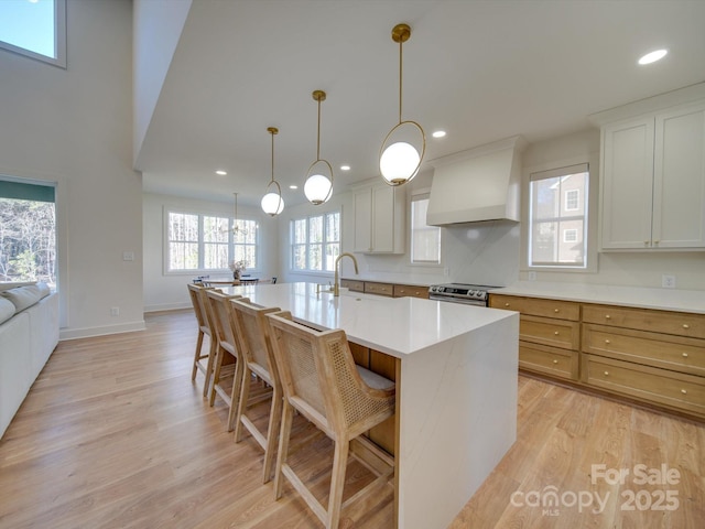 kitchen with premium range hood, white cabinetry, hanging light fixtures, a kitchen island with sink, and light hardwood / wood-style floors