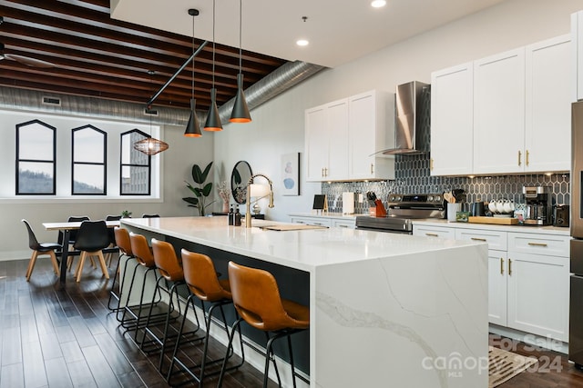 kitchen featuring white cabinetry, hanging light fixtures, wall chimney range hood, an island with sink, and stainless steel range with electric stovetop