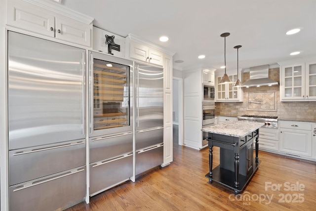 kitchen featuring wall chimney exhaust hood, built in appliances, a center island, hanging light fixtures, and white cabinets