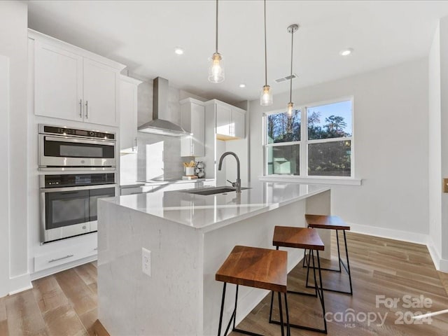 kitchen with a center island with sink, wall chimney range hood, sink, hanging light fixtures, and white cabinetry