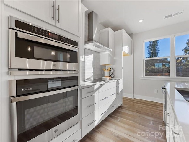 kitchen featuring white cabinets, wall chimney exhaust hood, tasteful backsplash, light hardwood / wood-style floors, and stainless steel double oven