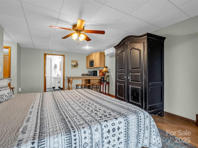 bedroom with a paneled ceiling, ensuite bathroom, a wall mounted AC, ceiling fan, and hardwood / wood-style flooring