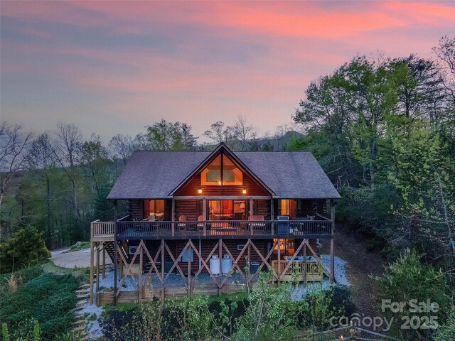 back of house at dusk featuring covered porch, roof with shingles, stairs, and log exterior