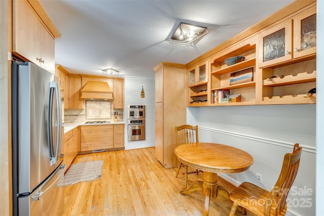 kitchen with light hardwood / wood-style flooring, decorative backsplash, light brown cabinetry, custom range hood, and appliances with stainless steel finishes