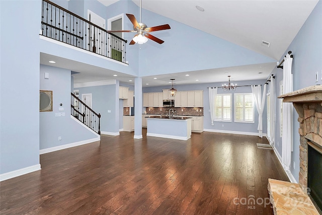 unfurnished living room featuring sink, dark wood-type flooring, high vaulted ceiling, a fireplace, and ceiling fan with notable chandelier