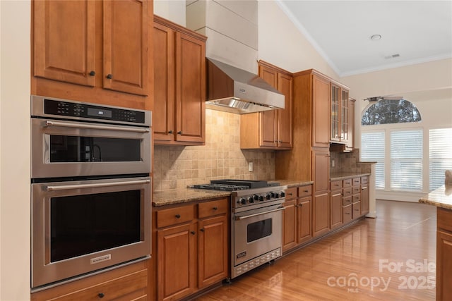 kitchen featuring crown molding, wall chimney exhaust hood, light hardwood / wood-style floors, light stone counters, and stainless steel appliances