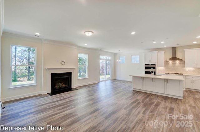 kitchen featuring white cabinetry, hanging light fixtures, a kitchen island with sink, decorative backsplash, and wall chimney range hood