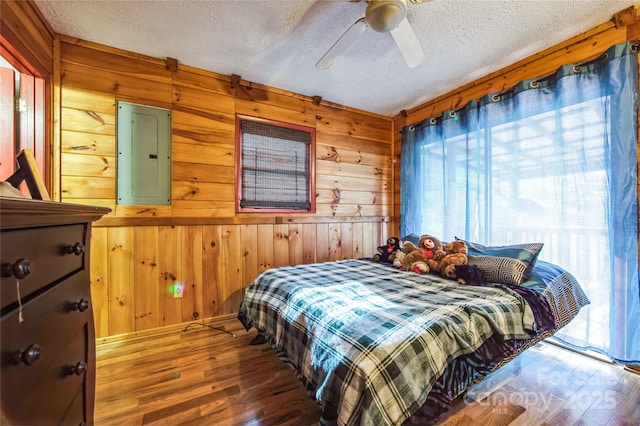 bedroom featuring hardwood / wood-style flooring, ceiling fan, wood walls, and electric panel