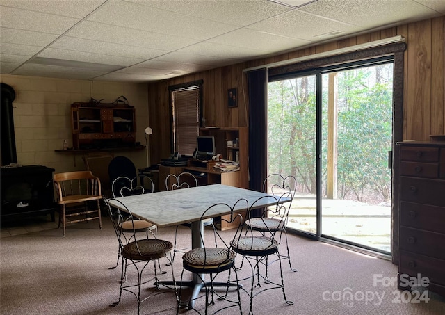 dining space featuring carpet flooring, a wood stove, a drop ceiling, and wooden walls