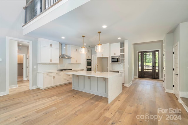 kitchen with white cabinets, an island with sink, decorative light fixtures, and wall chimney range hood