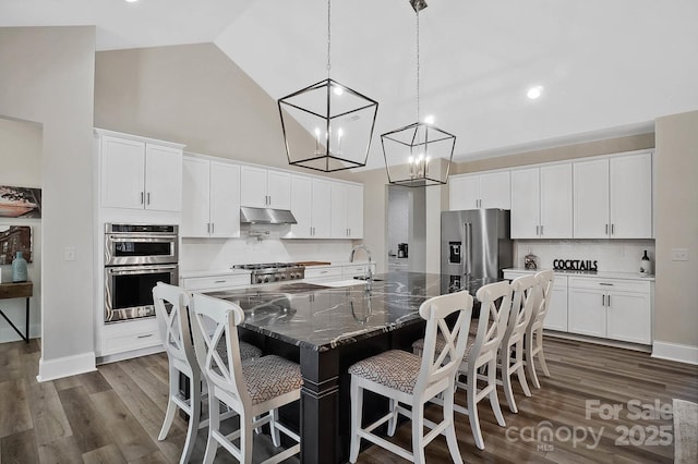 kitchen with white cabinetry, high vaulted ceiling, dark stone countertops, an island with sink, and appliances with stainless steel finishes