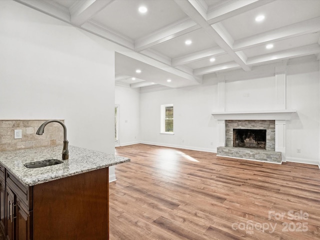 unfurnished living room featuring beam ceiling, sink, coffered ceiling, a stone fireplace, and light hardwood / wood-style flooring