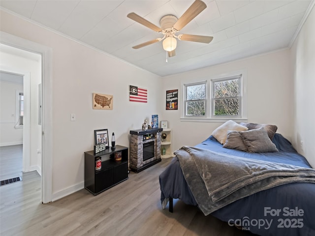 bedroom with light hardwood / wood-style floors, a stone fireplace, ceiling fan, and crown molding