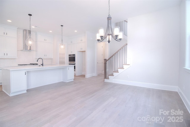 kitchen with light countertops, hanging light fixtures, a kitchen island with sink, white cabinetry, and wall chimney range hood