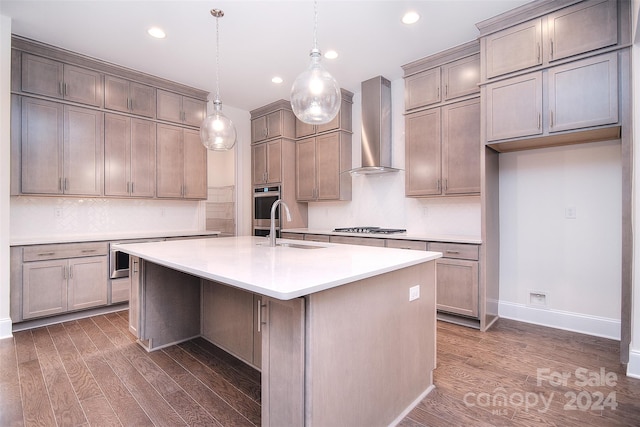 kitchen with dark wood-type flooring, sink, wall chimney range hood, pendant lighting, and a center island with sink