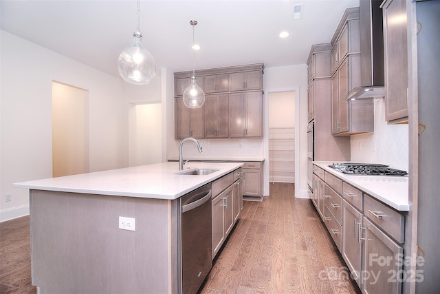 kitchen featuring a center island with sink, stainless steel appliances, a sink, wall chimney range hood, and wood finished floors