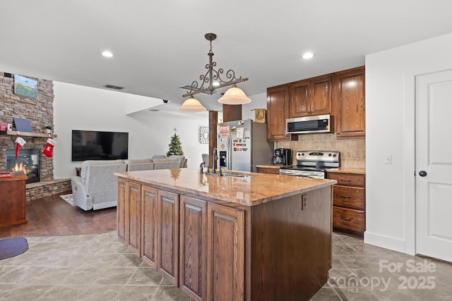 kitchen with sink, light stone counters, appliances with stainless steel finishes, an island with sink, and pendant lighting