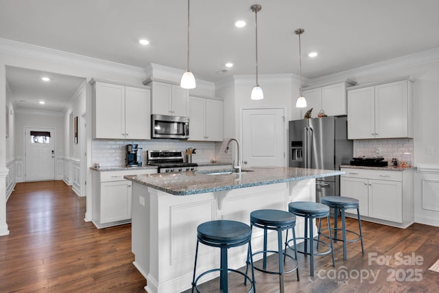 kitchen with appliances with stainless steel finishes, a kitchen island with sink, sink, white cabinets, and hanging light fixtures