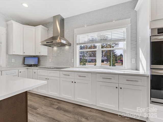 kitchen featuring decorative backsplash, black electric cooktop, double oven, wall chimney range hood, and white cabinetry