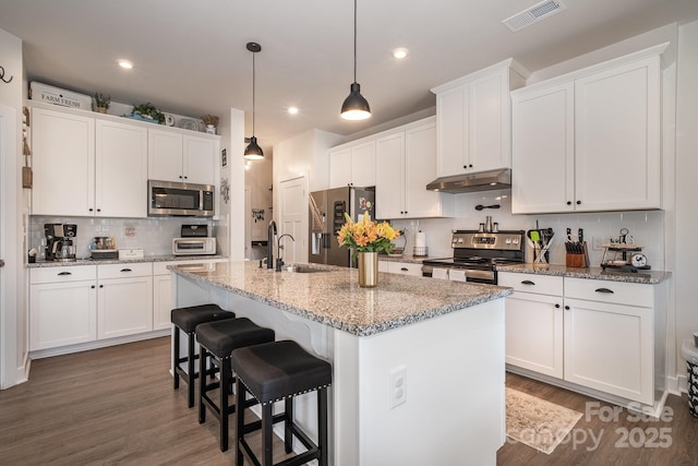 kitchen featuring a kitchen island with sink, white cabinets, light stone countertops, appliances with stainless steel finishes, and decorative light fixtures