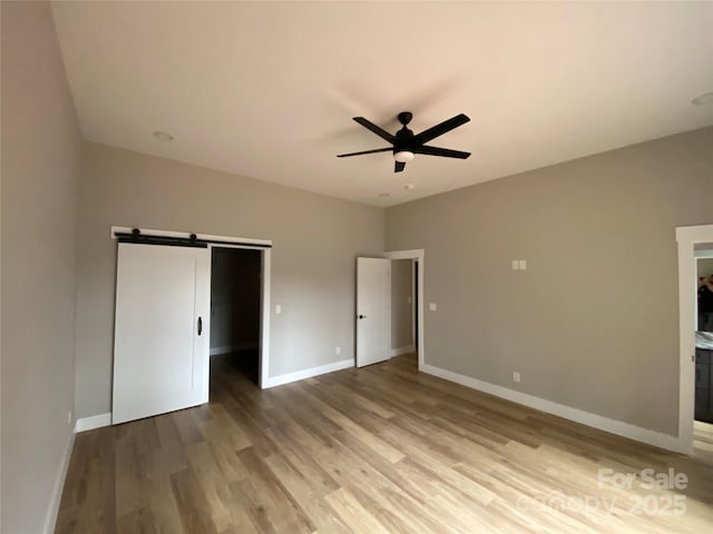 unfurnished bedroom with a walk in closet, ceiling fan, baseboards, a barn door, and wood finished floors