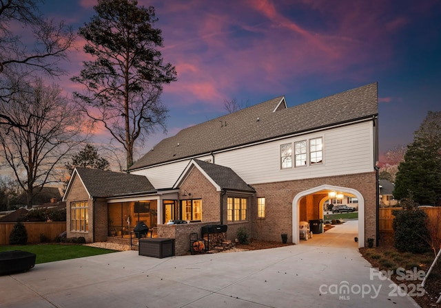 back of property at dusk with concrete driveway, fence, brick siding, and a patio