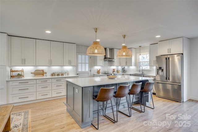 kitchen with high quality fridge, wall chimney exhaust hood, a kitchen island, white cabinetry, and hanging light fixtures