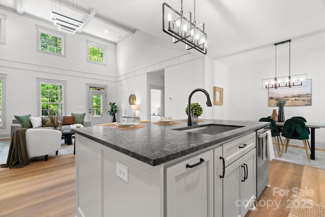 kitchen with light wood-type flooring, a kitchen island with sink, sink, decorative light fixtures, and white cabinets