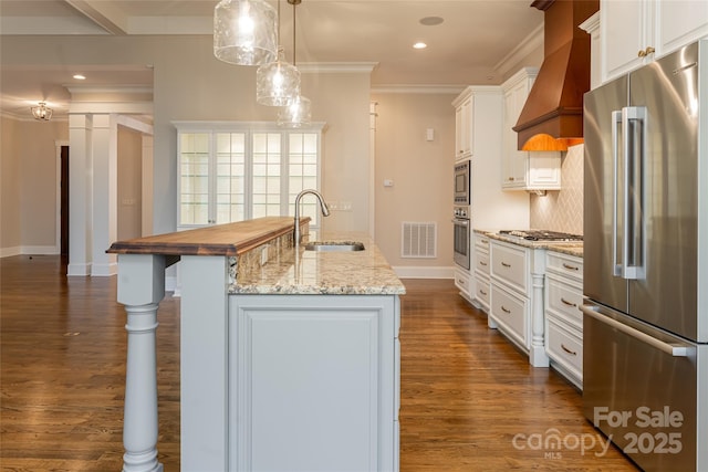 kitchen with a sink, stainless steel appliances, a center island with sink, premium range hood, and ornamental molding