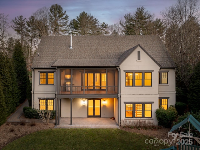 back of house at dusk featuring a lawn, a patio area, stucco siding, and a shingled roof