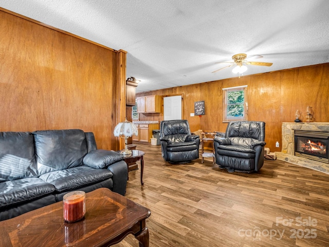 living room featuring a textured ceiling, ceiling fan, light hardwood / wood-style floors, a stone fireplace, and wood walls