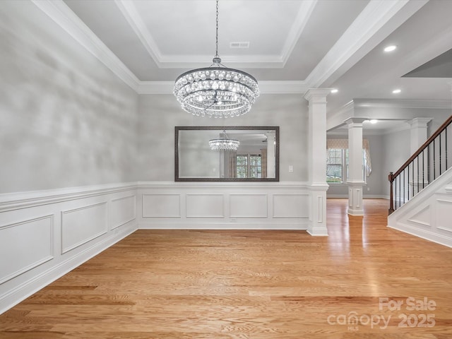 unfurnished dining area featuring stairway, an inviting chandelier, a tray ceiling, light wood-type flooring, and ornate columns