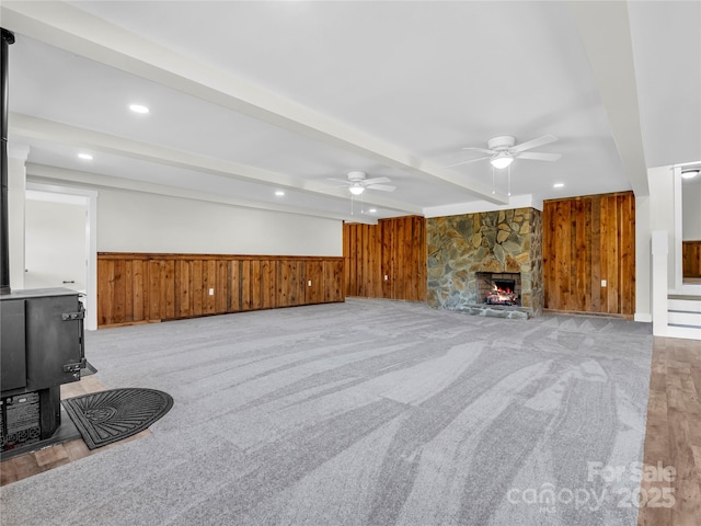 carpeted living room with ceiling fan, beam ceiling, a fireplace, and wooden walls