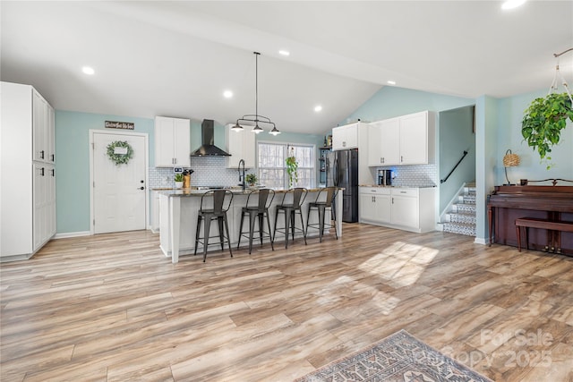 kitchen featuring a kitchen island with sink, wall chimney range hood, vaulted ceiling, white cabinetry, and stainless steel refrigerator