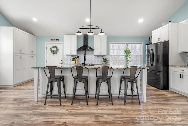 kitchen with wall chimney exhaust hood, white cabinetry, a center island with sink, and refrigerator