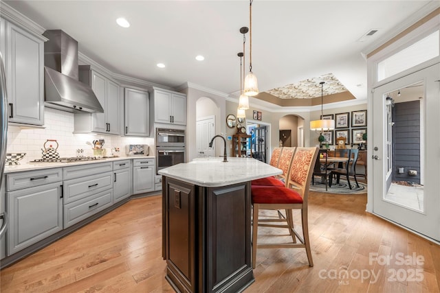 kitchen with wall chimney exhaust hood, light stone counters, light hardwood / wood-style flooring, an island with sink, and decorative light fixtures