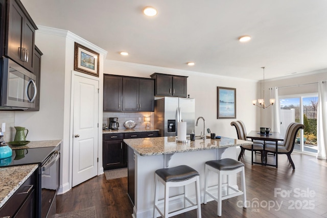 kitchen with light stone counters, dark hardwood / wood-style flooring, an island with sink, decorative light fixtures, and appliances with stainless steel finishes