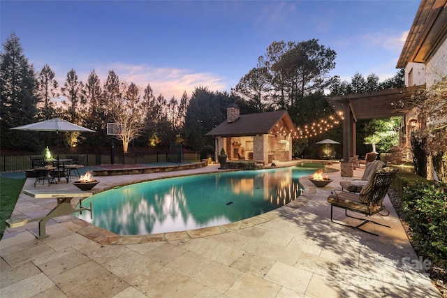 pool at dusk featuring a patio, an outbuilding, fence, an outdoor fire pit, and a storage structure