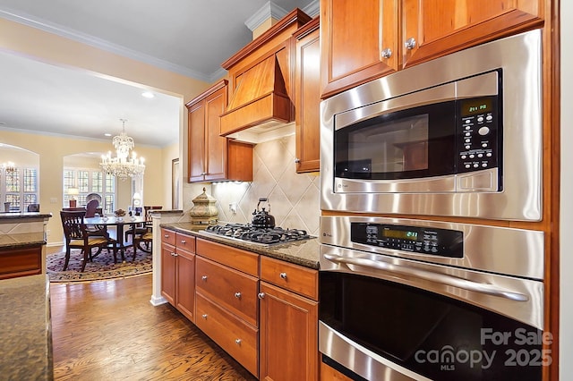 kitchen with decorative backsplash, ornamental molding, custom range hood, stainless steel appliances, and a notable chandelier