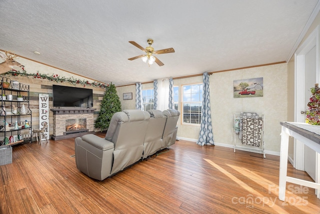 living room with a stone fireplace, a textured ceiling, wood-type flooring, ornamental molding, and ceiling fan