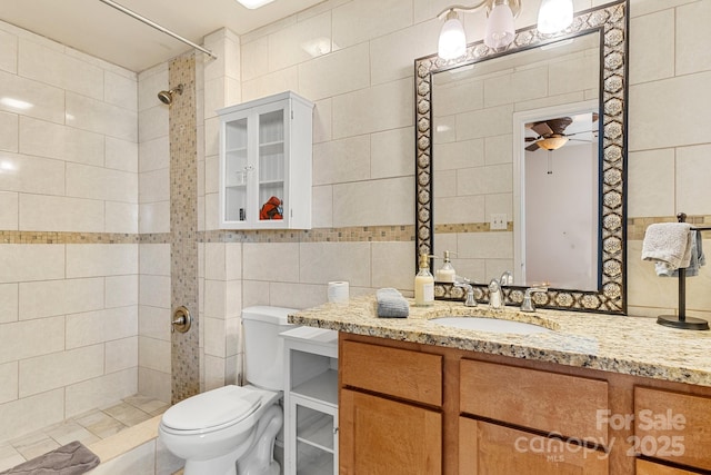 bathroom featuring ceiling fan, toilet, tiled shower, vanity, and tile walls