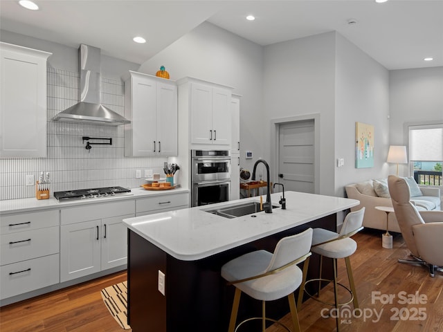 kitchen with stainless steel appliances, white cabinetry, a kitchen island with sink, and wall chimney range hood