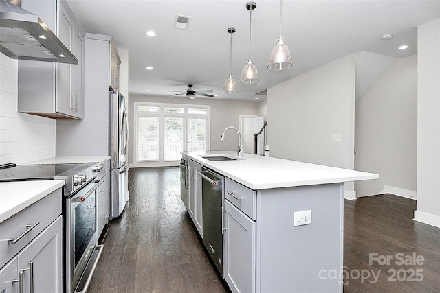 kitchen with appliances with stainless steel finishes, sink, wall chimney range hood, decorative light fixtures, and an island with sink