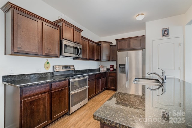 kitchen with sink, stainless steel appliances, dark stone countertops, dark brown cabinets, and light wood-type flooring