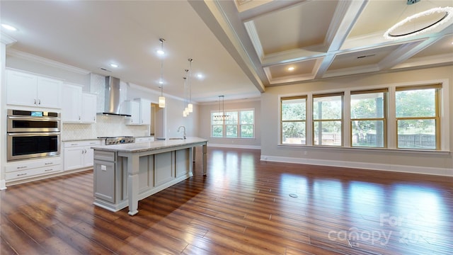 kitchen featuring light stone countertops, wall chimney exhaust hood, a kitchen island with sink, decorative light fixtures, and white cabinetry