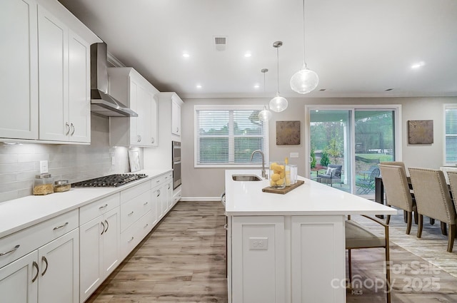 kitchen with decorative light fixtures, sink, appliances with stainless steel finishes, an island with sink, and wall chimney exhaust hood